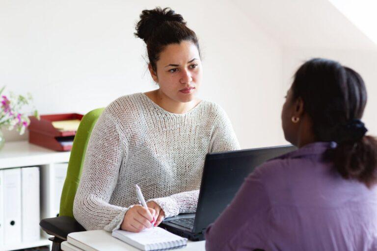 Two people seated at a desk during a support or advice session, with one person taking notes while speaking with the other across a laptop in an office setting.