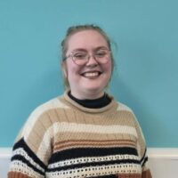 Head and shoulders portrait short of smmiling person with glasses, blonde hair and a brown striped jumper