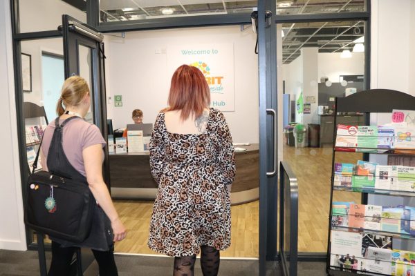 Two people walking into an office, In the background there is a reception desk with someone sat behind it. There is a leaflet stand to the righ of the image.