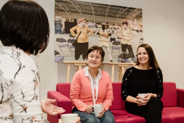 Two people sat on a pink sofe, there is a large picture behind them. One is wearing peach and the other is wearing black. Both are holding cups and are engaged in conversation with someone in the foreground with their back to the camera.